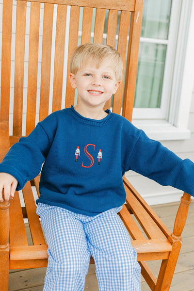 Child wearing a navy sweater with red letter 'S' monogram, sitting on a wooden chair.
