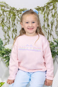 Child wearing a pink sweatshirt with rainbow applique, sitting against a white background with greenery.