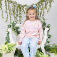 girl in a pink roll neck sweater with custom applique and light blue pants sitting on a white chair surrounded by greenery.