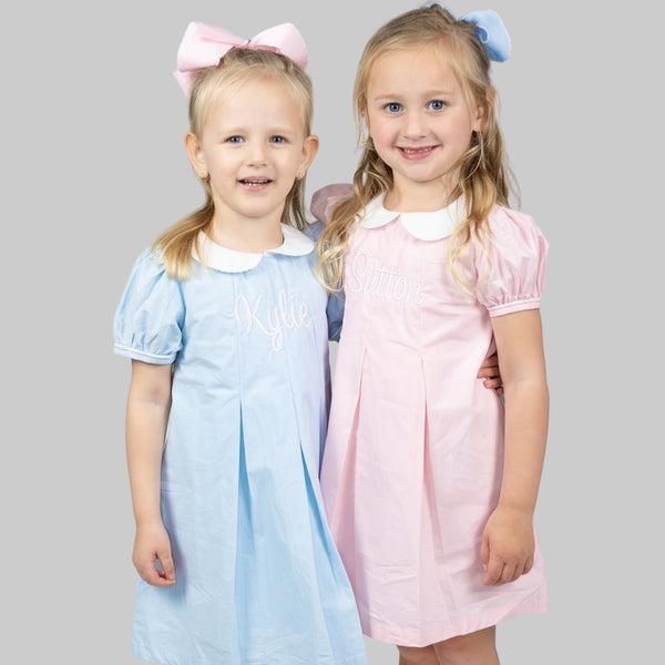 Two girls standing next to each other, wearing dresses with Peter Pan collars. One dress is blue and the other is pink.